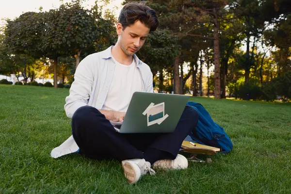 Genç bir adam parkta çimlerde oturur, laptopunu çalışmak ya da çalışmak için kullanır. Doğa ile çevrili bu sakin açık havada verimliliği ve rahatlığı somutlaştırıyor..
