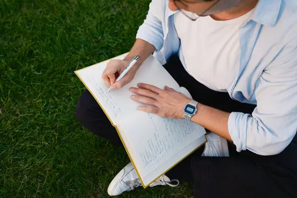 Çimlerin üzerinde oturan bir kişi dışarıda kalemle yazarken bir defter tutar. Ortam, sakin bir doğal ortamın altında yansıtıcı ya da yaratıcı bir an olduğunu gösteriyor..