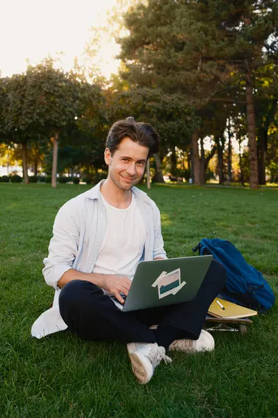 Parktaki çimlerin üzerinde dizüstü bilgisayarla oturan bir adam dışarıda çalışır ya da ders çalışır. Rahatlamış görünüyor, çevresi yeşilliklerle çevrili ve doğal bir ortamın tadını çıkarıyor..