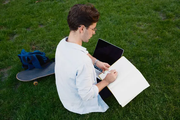 Çimenli bir arazide oturan genç bir adam, bir dizüstü bilgisayar ve kaykay ve sırt çantasıyla ders çalışıyor. Sıradan, akademik ve rahatlatıcı bir atmosfer yakalar..