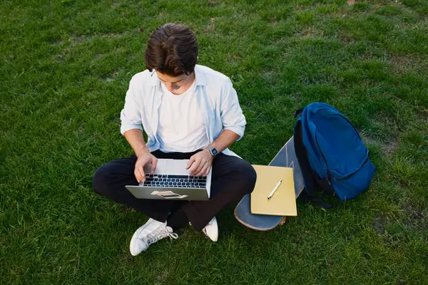 Çimlerin üzerinde oturan genç bir adam, yanında bir kaykay ve sırt çantası ile bir dizüstü bilgisayarla çalışıyor ya da çalışıyor..
