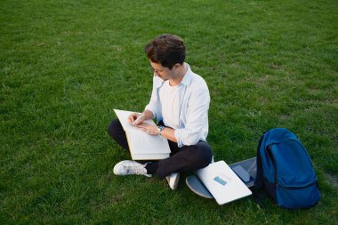 Bir not defterine yazarken parkta çimenlikte oturan genç bir adam, bir dizüstü bilgisayar, bir sırt çantası ve bir kaykay gibi çalışma materyalleriyle çevrili..
