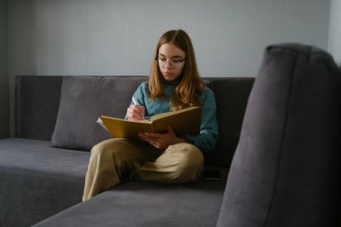 Genç kız, modern gri bir koltukta otururken deftere notlar yazıyor..