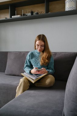 Genç bir kız dizüstü bilgisayar okurken akıllı telefon kullanarak kanepede oturdu.