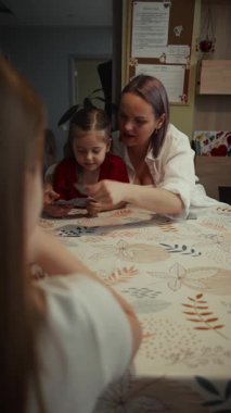 Bir aile mutfak masasında iskambil oynar. Anne kızının kağıt oynamasına yardım ediyor..