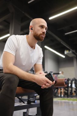 Akıllı telefon tutan, kulaklık takan, spor salonunda spor aletleriyle oturan bir adam.