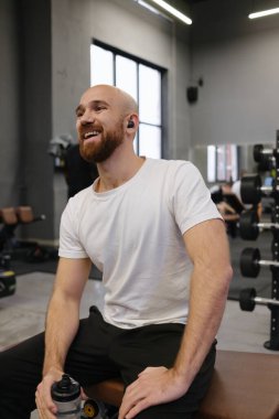 Gülümseyen bir adam spor salonunda oturur, egzersiz sonrası memnuniyeti ve coşkuyu gösterir..
