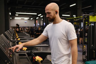 Odaklanmış adam egzersiz ekipmanlarıyla dolu iyi aydınlatılmış bir spor salonunda koşu bandı kullanıyor.