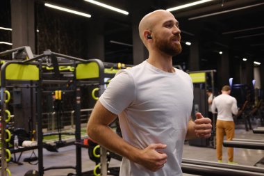 Arka planda egzersiz ekipmanıyla spor salonunda koşan bir adam.