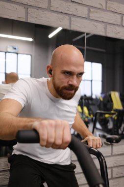 Beyaz tişörtlü bir adam spor salonunda bisiklet sürüyor..