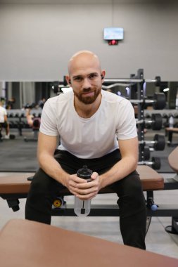 Egzersiz sırasında mola veren bir adam, spor salonunda su şişesiyle bankta oturuyor.