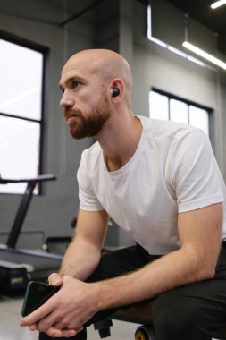 Spor salonunda oturan kel sakallı adam elinde cep telefonu ve kulaklık tutuyordu..
