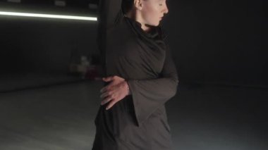 Siyah elbiseli bir kadın duygusal hareketlerle karanlık bir stüdyoda dans ediyor..