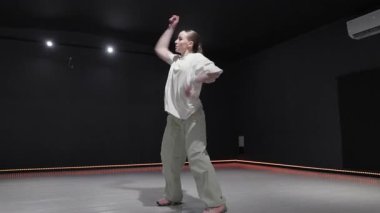 Modern bir stüdyoda eşsiz ışıklandırmayla dans eden genç bir kadın. O rahat ve şık kıyafetler giyiyor. Dans stili çağdaş..