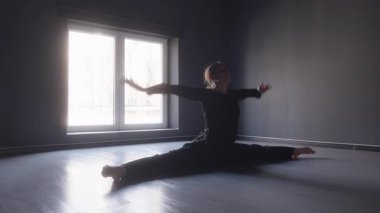 Siyah elbiseli bir dansçı, büyük pencereli karanlık bir odada ön kapıyı açar..