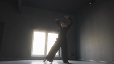 Siyah kıyafetli, aydınlık pencereli karanlık bir odada çağdaş dans eden bir kadın..