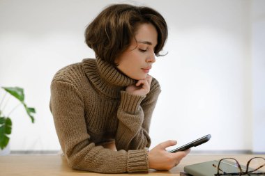 Kışlık süveter giyen genç bir kadın kapalı bir masada akıllı telefon kullanıyor.