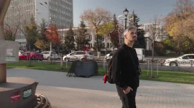 Kameralı ve sırt çantalı bir adam canlı bir şehir parkında keyifli bir yürüyüş yapıyor..