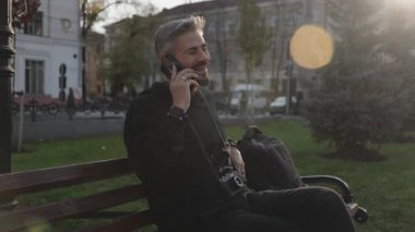 Memnun adam güneşli bir park ortamında cep telefonuyla neşeli bir sohbetten hoşlanır..
