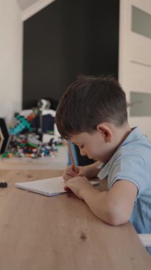 Genç bir çocuk ahşap bir masada oturuyor, özenle bir deftere çizim yapıyor. Bu video çocukluk, yaratıcılık ve öğrenmeyi çağrıştırıyor..