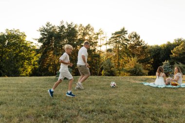 Bir aile dışarıda futbol oynayıp piknik battaniyesinde dinlenir..