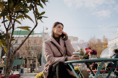 A woman sits at a cafe table, talking on her phone, with a coffee cup in hand and a pink quilted jacket outdoors.