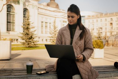 Professional woman on a bench outdoors using a laptop in a city square, coffee nearby.