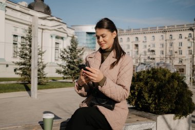 A woman in a pink coat sits on a bench outdoors, texting on her smartphone with a coffee cup nearby.