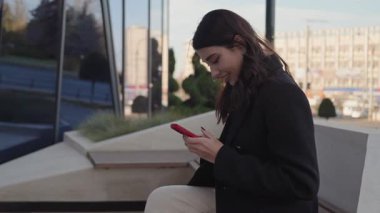 Genç bir kadın, şehir bankında cep telefonuyla iletişim kurarken ışıl ışıl gülümsüyor..