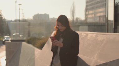 Cep telefonuna dalmış, odaklanmış bir kadın, çağdaş bir şehir ortamında dışarıda duruyor..