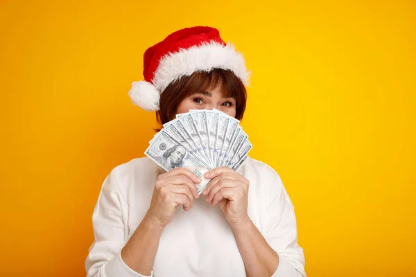 感情的な幸せな成熟した中年の女性で赤いサンタクロースの帽子黄色の背景にお金を保持する クリスマスビジネスのコンセプト ストック写真