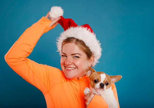 青の背景に隔離されたChihuahua犬とオレンジのTシャツで幸せな若いサンタクラスの女性 人々クリスマスライフスタイルの休日のコンセプト ストック画像