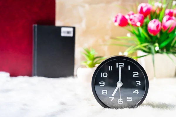 Black alarm clock and book with pink flower on table