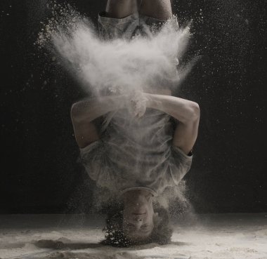 Spor kıyafetli genç bir erkek dansçı karanlık bir odada beyaz toz bulutu içinde amuda kalkıyor.