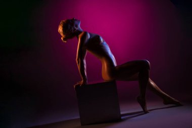 Stüdyoda gümüş küpün üzerinde renkli ışıkla oturan çıplak bir kadın.