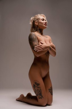 Vücudunda sarı saç ve dövmeler olan şehvetli bir kadın model stüdyoda gri arka planda oturuyor.