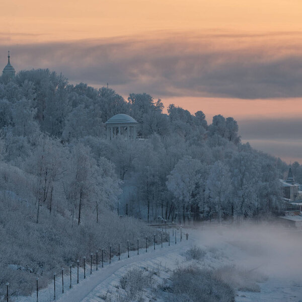 Beautiful Russian winter landscape with frozen river, embankment, street lamps and white rotund on a hill against pink and yellow sunset view