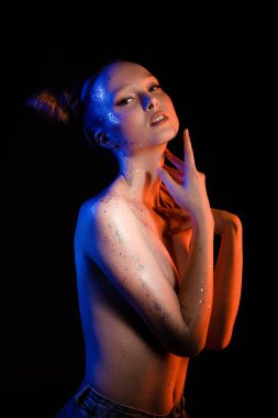 Kendine güvenen, kendine güvenen, yaratıcı saç stili ve göğsünü kapatan ve siyah arka planda kameraya bakan genç kadın modelin yan görüntüsü.