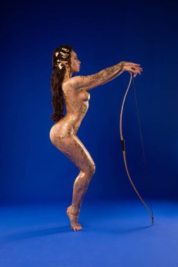 Çıplak vücudunda altın rengi boya, oklu elf kulakları ve mavi arka planda yay dansı olan kendinden emin kadın manken.