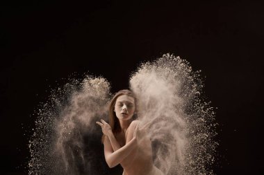 İç çamaşırı giymiş, gözlerini kapayan koyu arkaplanda kum sıçrayan tam boy çıplak genç bir kadın.