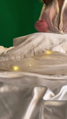 Beyaz elbiseli anonim bir kadın fotoğraf çekimi sırasında parlak çelenklerle aydınlık yatak odasında rahat bir yatakta oturuyor.