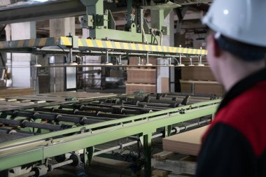 Üniformalı bir fabrika işçisinin endüstriyel taşıyıcının yanında dikilip fabrikada çalışırken modern mobilya detaylarını üretmesi.