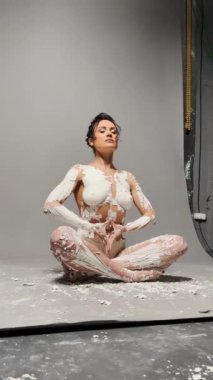 Bir kadın stüdyo ortamında yoga pozisyonunda oturuyor. Tüm vücudu beyaz boyayla kaplı. Boya lekelenmiş, dağınık ve sanatsal bir görünüm yaratıyor. Arkaplan basit bir gri fon.