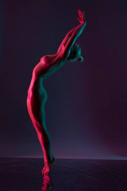 Sarışın, sırtüstü uzanmış, kollarını kaldırmış, koyu mor arka planda zarif dans eden genç bir kadın.