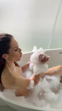 Lüks banyoda köpük banyosu yaparken küvette baloncuk çıkaran neşeli kadın görüntüsü