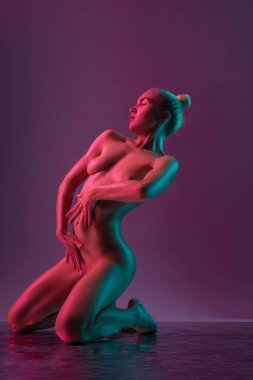 Stüdyoda dizlerinin üzerinde mor ışıkla yoga yapan üstsüz, şehvetli bir kadının yan görüntüsü.