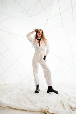 Beyaz tulumlu genç bir kadın beyaz bir odada poz veriyor yere beyaz kumaş örtülmüş. Onun saçı uzun ve kırmızı, ve o siyah eldiven giyiyor. Kumaş dokulu bir arkaplan oluşturur