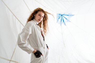 Kıvırcık saçlı genç bir kadın beyaz tulumlu ve siyah eldivenli beyaz bir stüdyoda poz veriyor. Kendine güvenen bir ifadesi var ve doğrudan kameraya bakıyor. Arka planda mavi bir kağıt var.