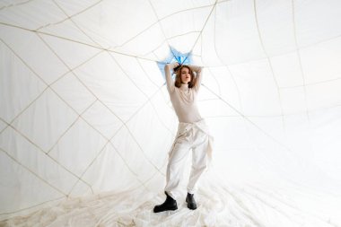 Beyaz elbiseli ve siyah çizmeli genç bir kadın ellerinde saçlarıyla beyaz duvarlar ve tavana gerilmiş beyaz bir kumaşla poz veriyor.