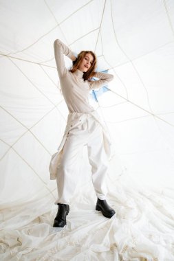 Beyaz elbiseli ve siyah çizmeli genç bir kadın ellerinde saçlarıyla beyaz duvarlar ve tavana gerilmiş beyaz bir kumaşla poz veriyor.
